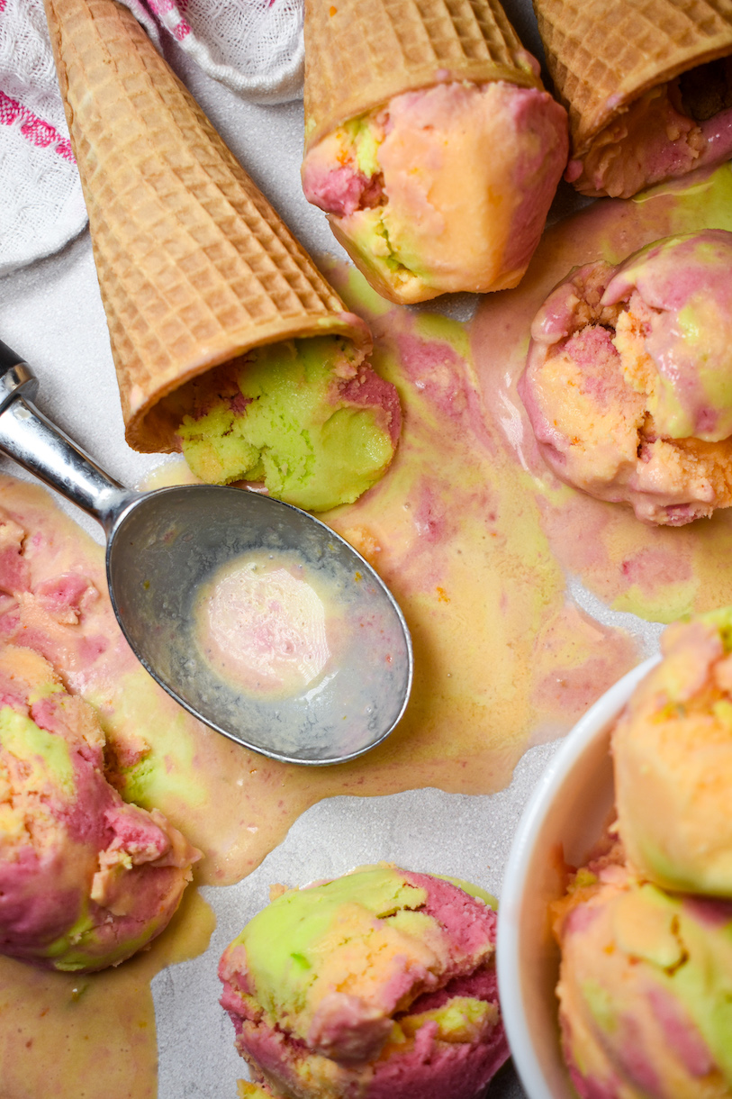 Ice cream cones, a metal ice cream scoop, and melted rainbow sherbet on a white surface