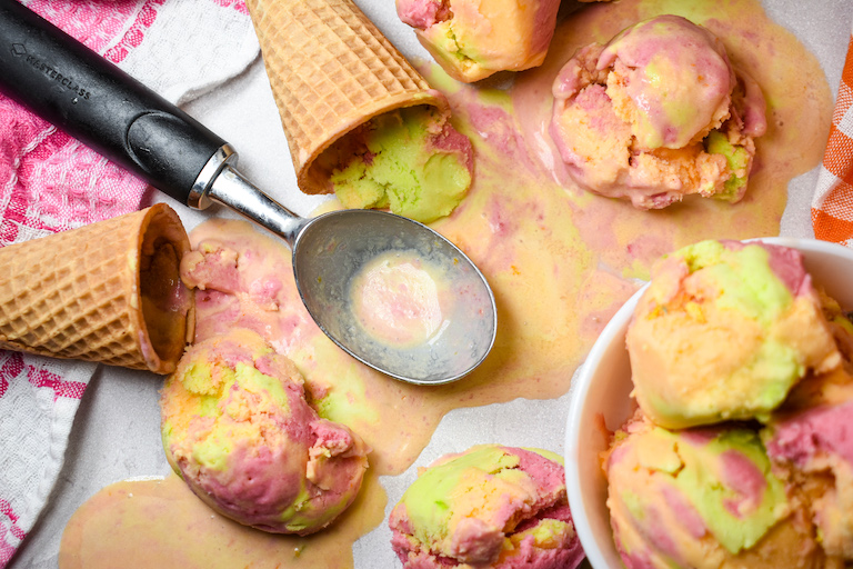 Melted rainbow sherbet, an ice cream scoop, and a bowl of sherbet on a white surface with sugar cones