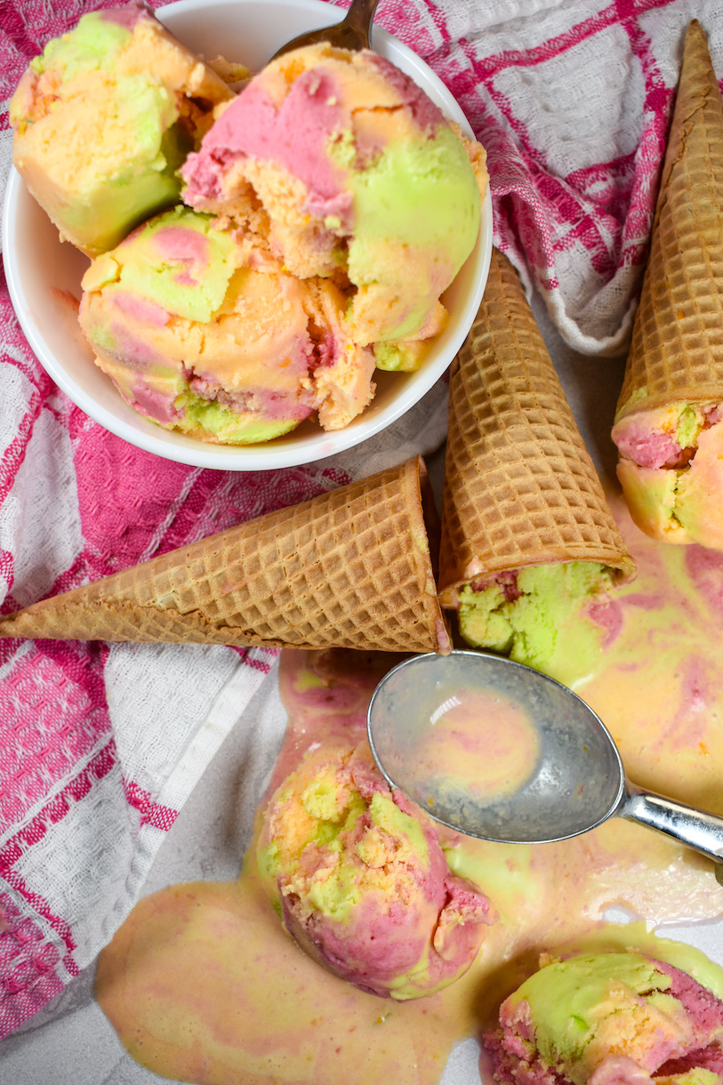 A bowl of rainbow sherbet, pink tea towel, ice cream cones, and ice cream scoop on a white surface