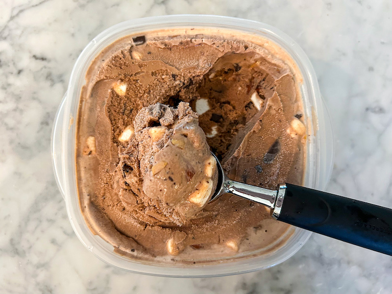 An ice cream scoop scooping rocky road ice cream from a plastic container