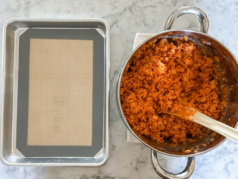 A small tray and a stock pot of orange cereal treat mixture arranged on a marble surface