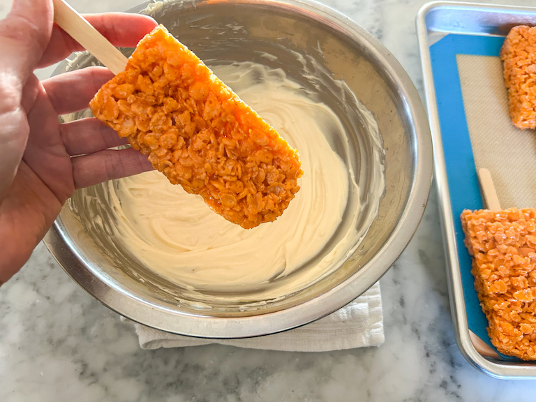 Hand dipping a Krispie treat pop in melted white chocolate