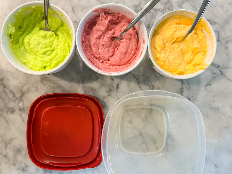 A plastic container and bowls of green, pink, and orange sherbet arranged on a marble surface