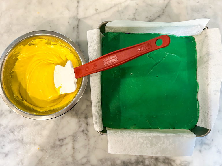 A bowl of yellow melted chocolate and a pan of green fudge on a marble surface