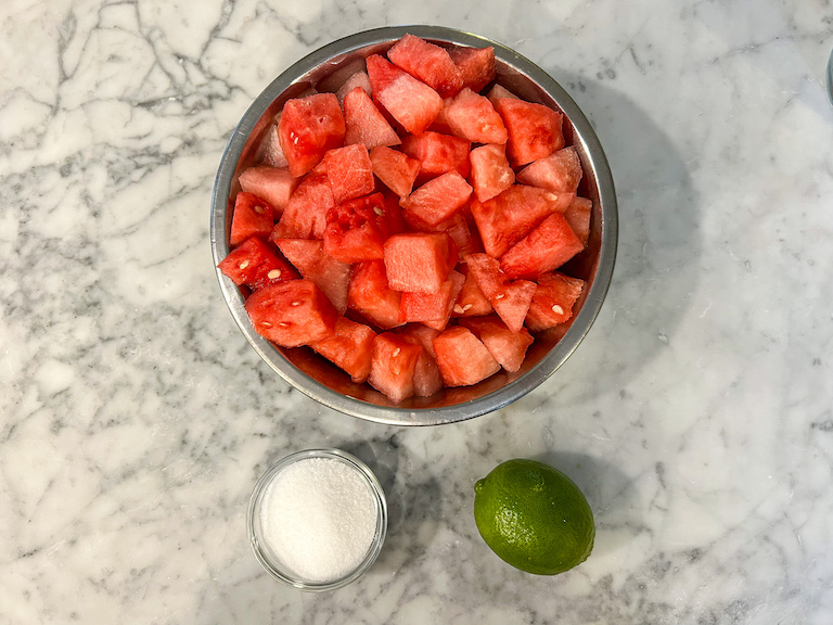Bowl of watermelon, bowl of sugar, and a lime positioned on a marble countertop