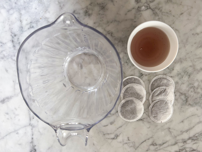A pitcher, six tea bags, and a dish of lavender syrup on a marble surface
