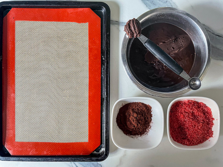 A small tray, dish of ganache, melon baller, and dishes of cocoa powder and red sugar on a marble surface