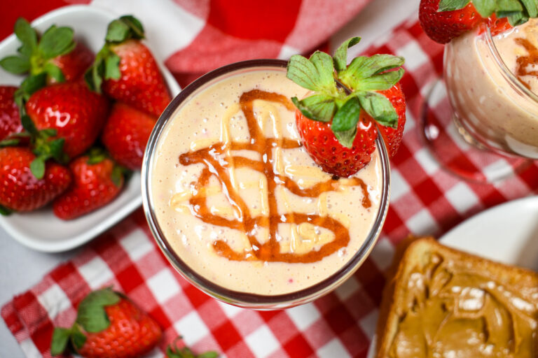 Looking down at a peanut butter and jelly smoothie positioned on a checkered tea towel with a dish of strawberries