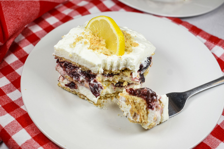 A slice of cherry cheesecake icebox cake garnished with a lemon slice, on a plate with a fork