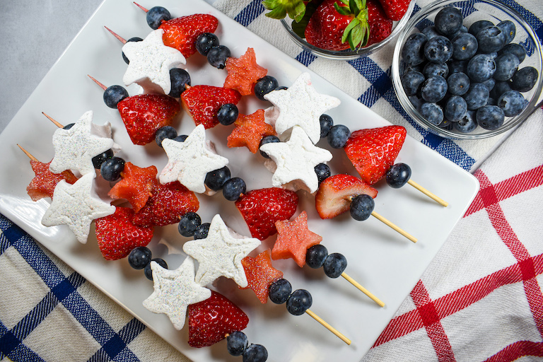 A platter of fruit skewers with dishes of fresh strawberries and blueberries arranged on plaid red and blue tea towels