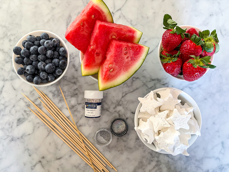 Dishes of blueberries, watermelon, strawberries, and marshmallows on a marble surface with wooden skewers