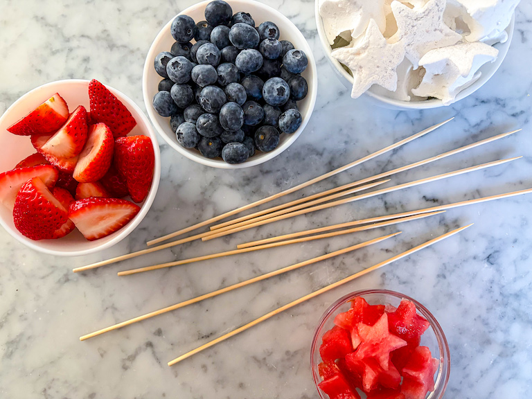 Dishes of berries, watermelon, and marshmallows on a marble surface with wooden skewers