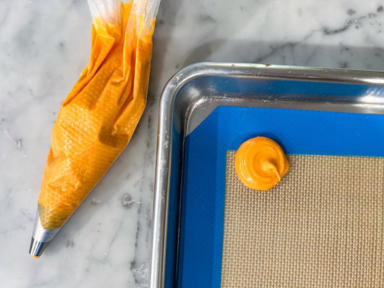 A piping bag of orange chocolate and a silicone lined tray piped with a dollop of orange chocolate
