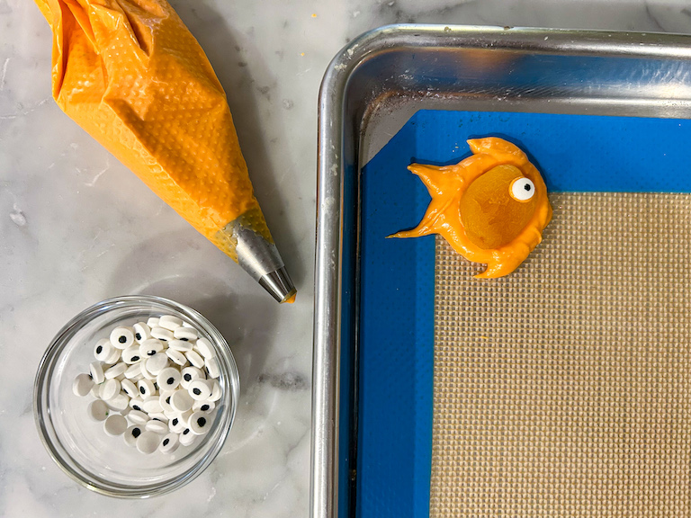 A goldfish piped on a silicone mat next to a piping bag and dish of candy eyeballs
