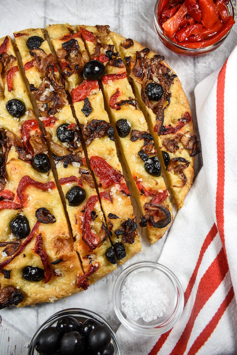 Loaf of focaccia bread with olives, caramelized onions, and peppers, next to a tea towel and dishes of salt and roasted red peppers