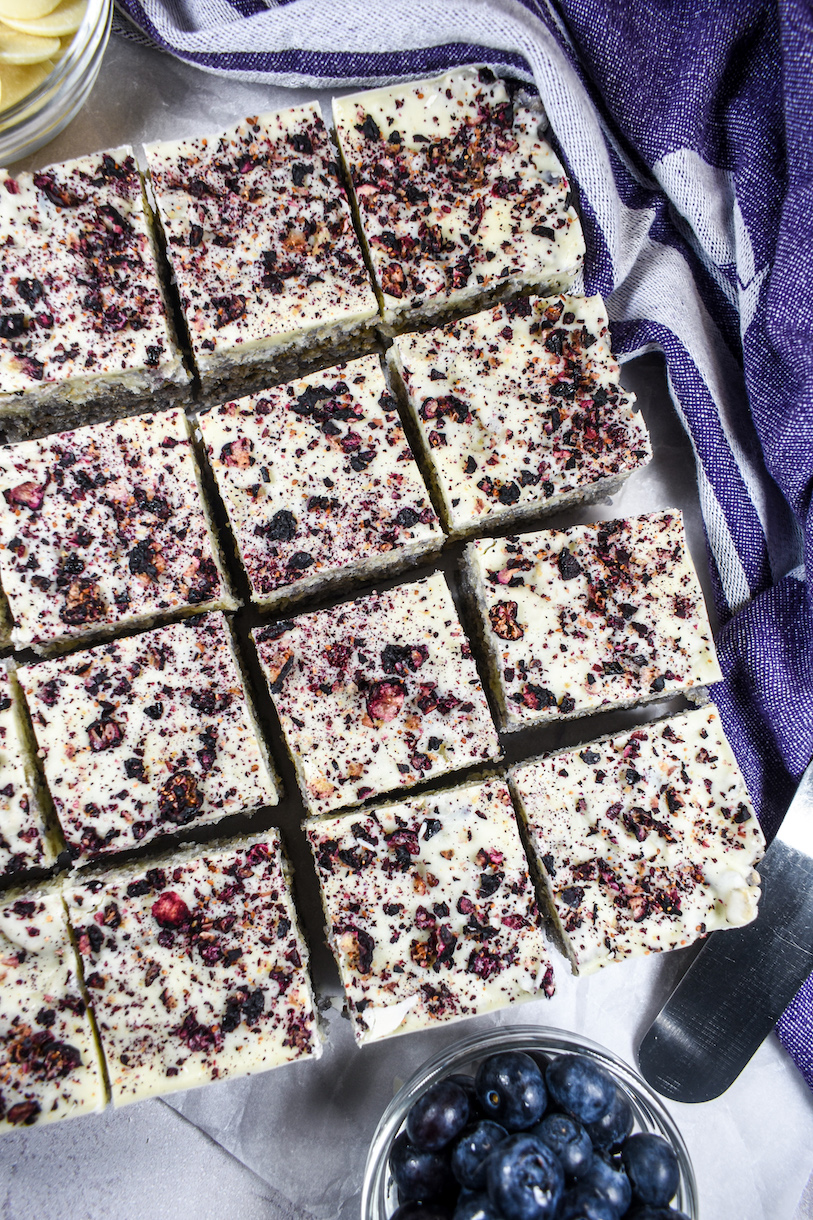 Slices of blueberry cereal treat on a white surface with a purple towel and a dish of blueberries