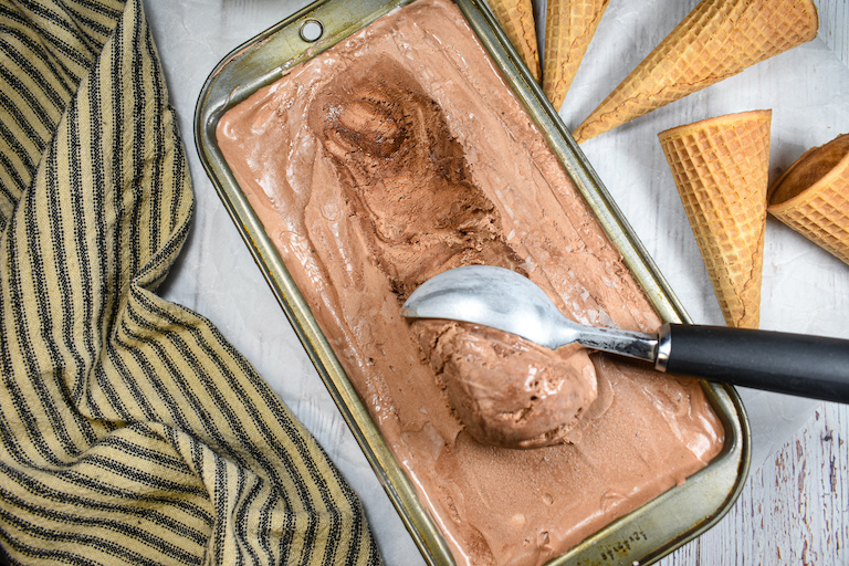 A loaf pan of no churn chocolate ice cream surrounded by cones, a striped towel, and an ice cream scoop