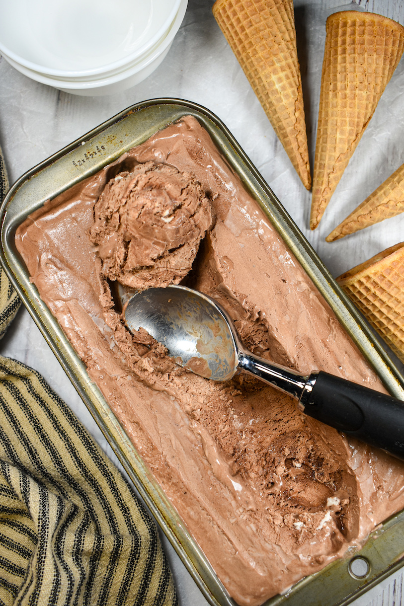 Vertical shot of a loaf pan of chocolate ice cream with a scoop, surrounded by ice cream cones, bowls, and a striped towel