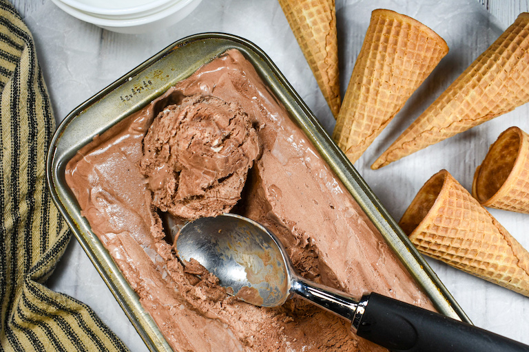 loaf pan of chocolate ice cream, ice cream scoop, cones, and striped tea towel on a white surface