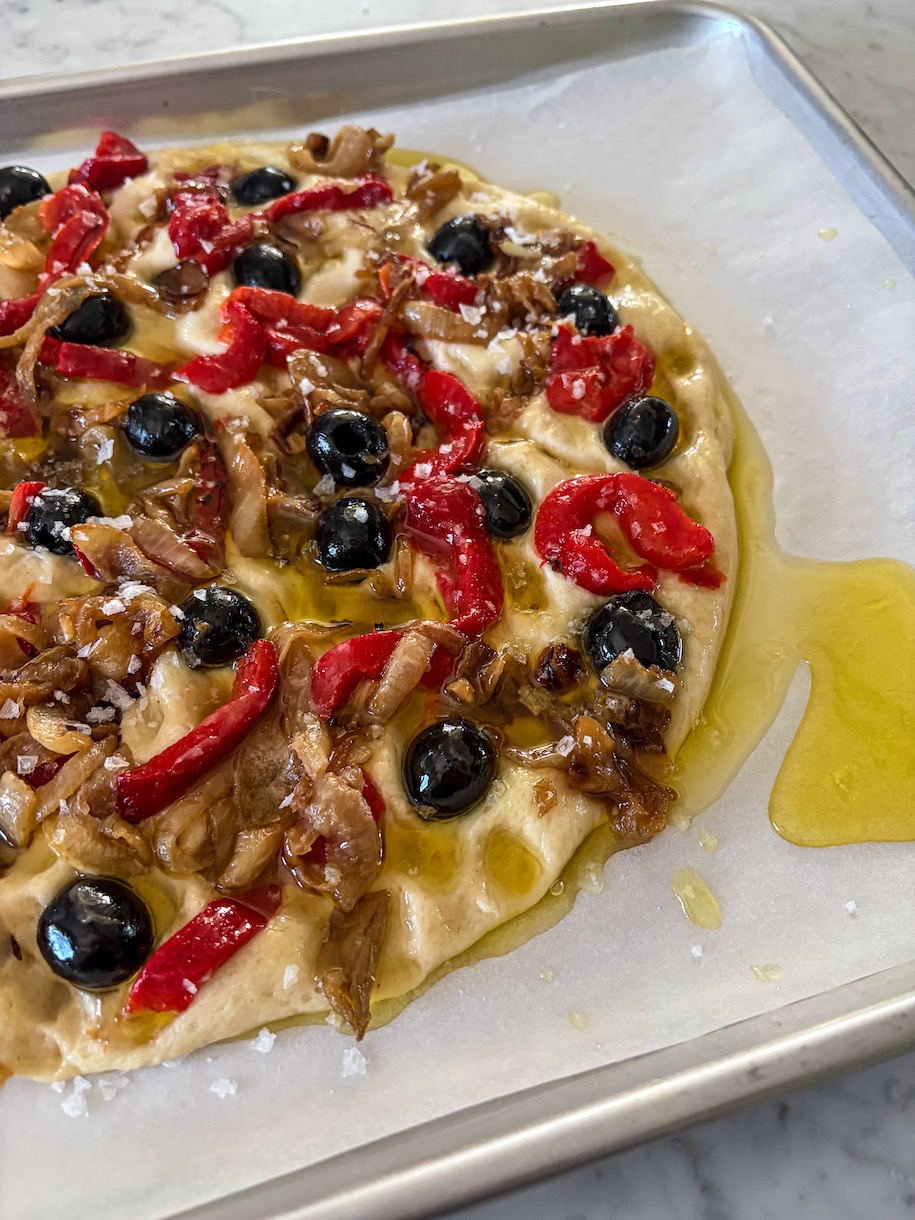 Unbaked focaccia topped with olives, onions, and peppers, drizzled with olive oil