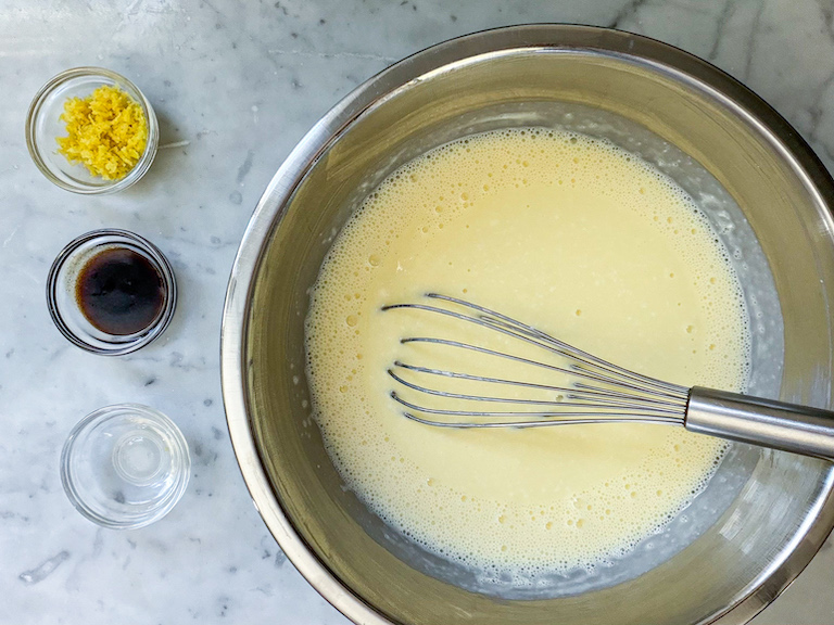 Ice cream base in a bowl with a whisk, next to dishes of lemon zest, vanilla, and vodka