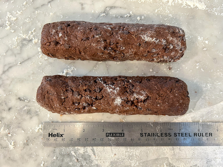 Two logs of chocolate biscotti dough on a marble surface with a ruler