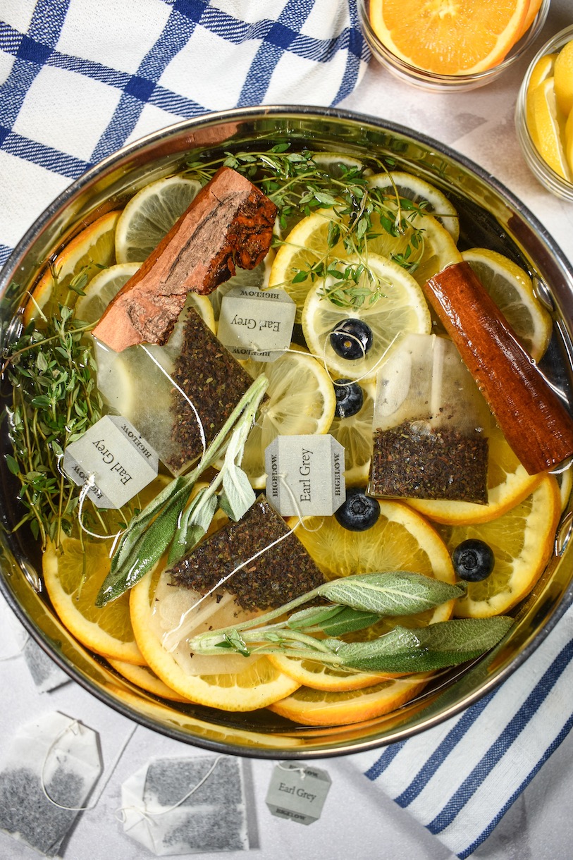 Looking down at a simmer pot with tea bags, citrus fruit, cinnamon sticks, and fresh herbs