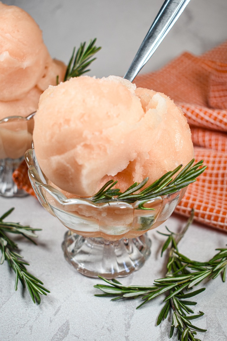 A glass dish of grapefruit and rosemary sorbet surrounded by rosemary sprigs