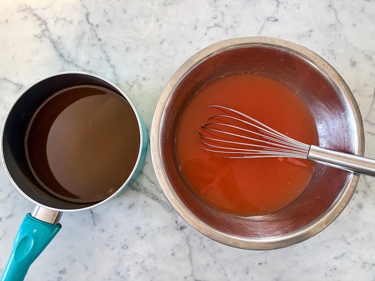Pan of grapefruit juice and bowl of grapefruit juice with a whisk, arranged on a marble surface