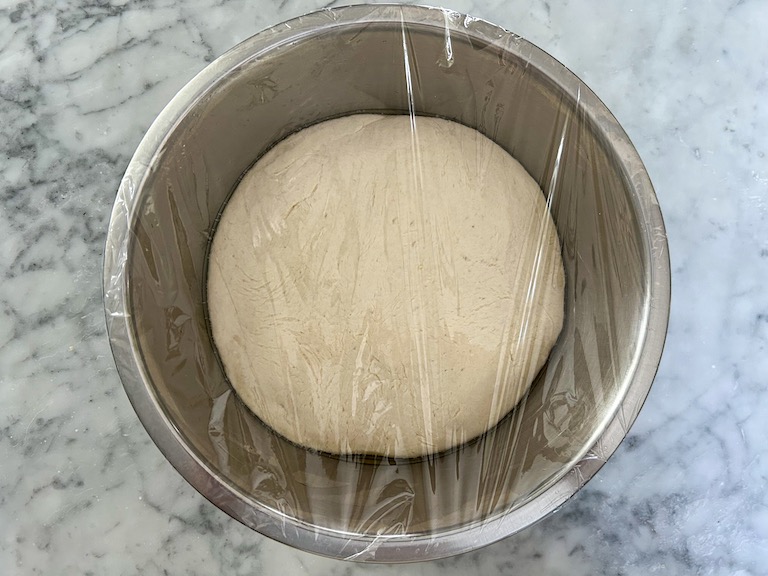 A bowl of pizza dough with plastic wrap stretched over the top