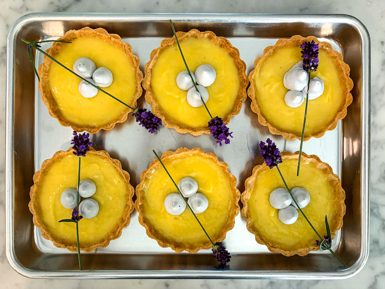 six mini lemon tarts on a tray, garnished with lavender whipped cream and fresh lavender flowers
