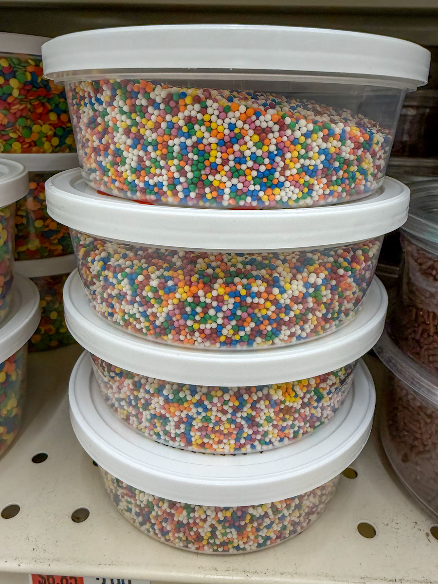 A stack of containers of rainbow nonpareil sprinkles on a shelf in a supermarket