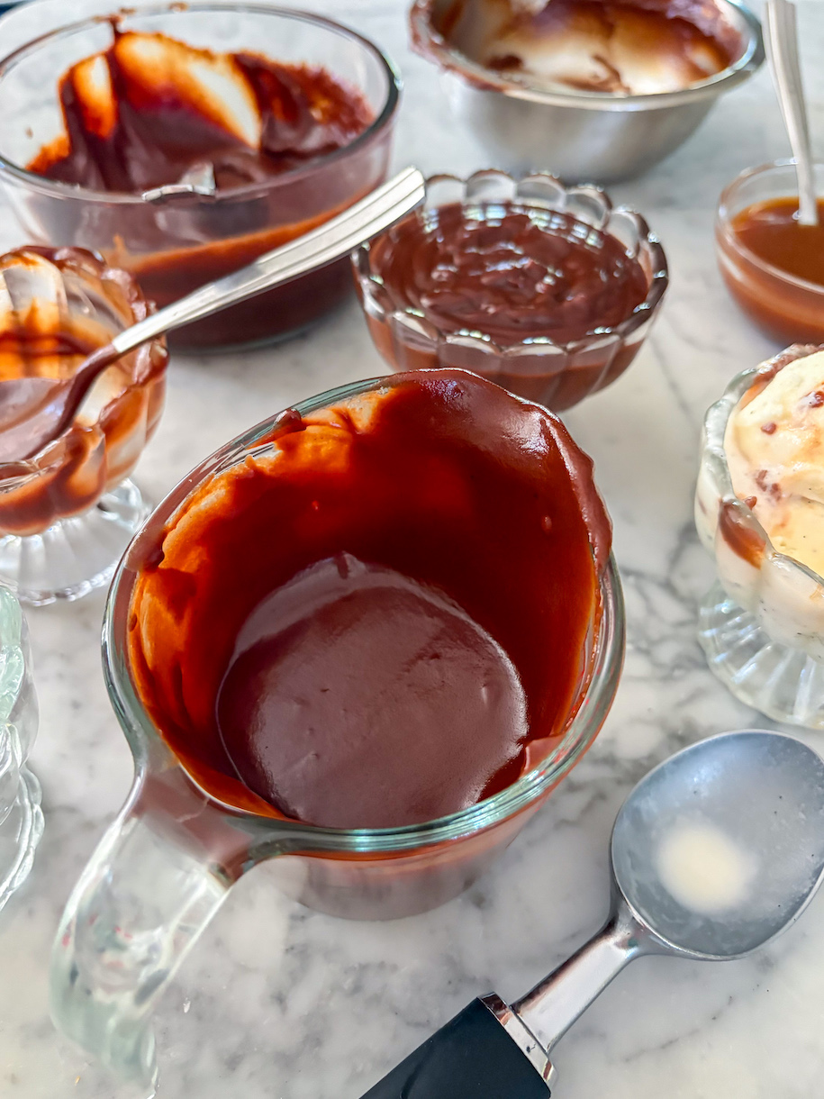 Cups and dishes of hot fudge sauce and dishes of ice cream scattered across a marble surface