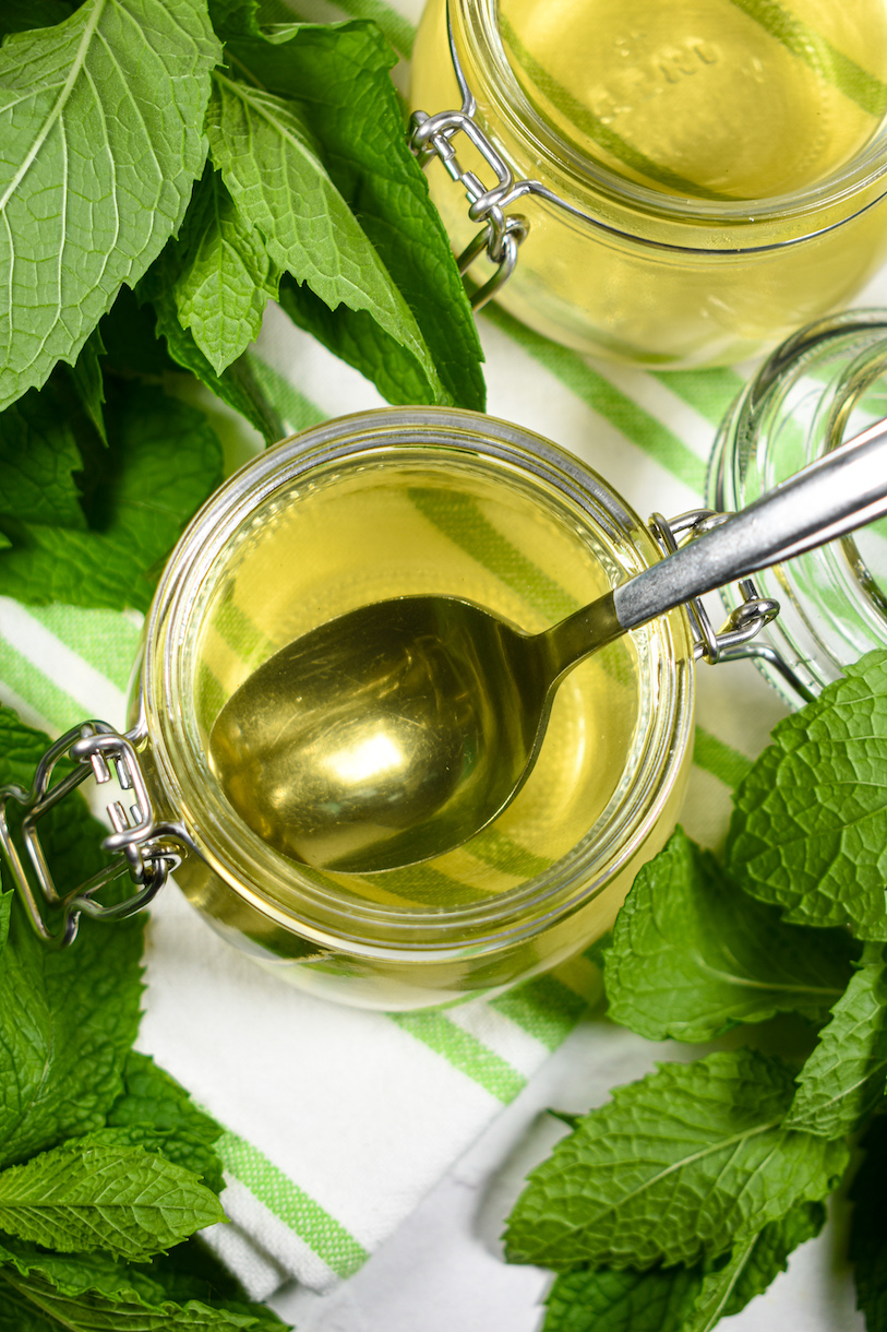 A jar of mint simple syrup with a spoon, surrounded by green mint leaves