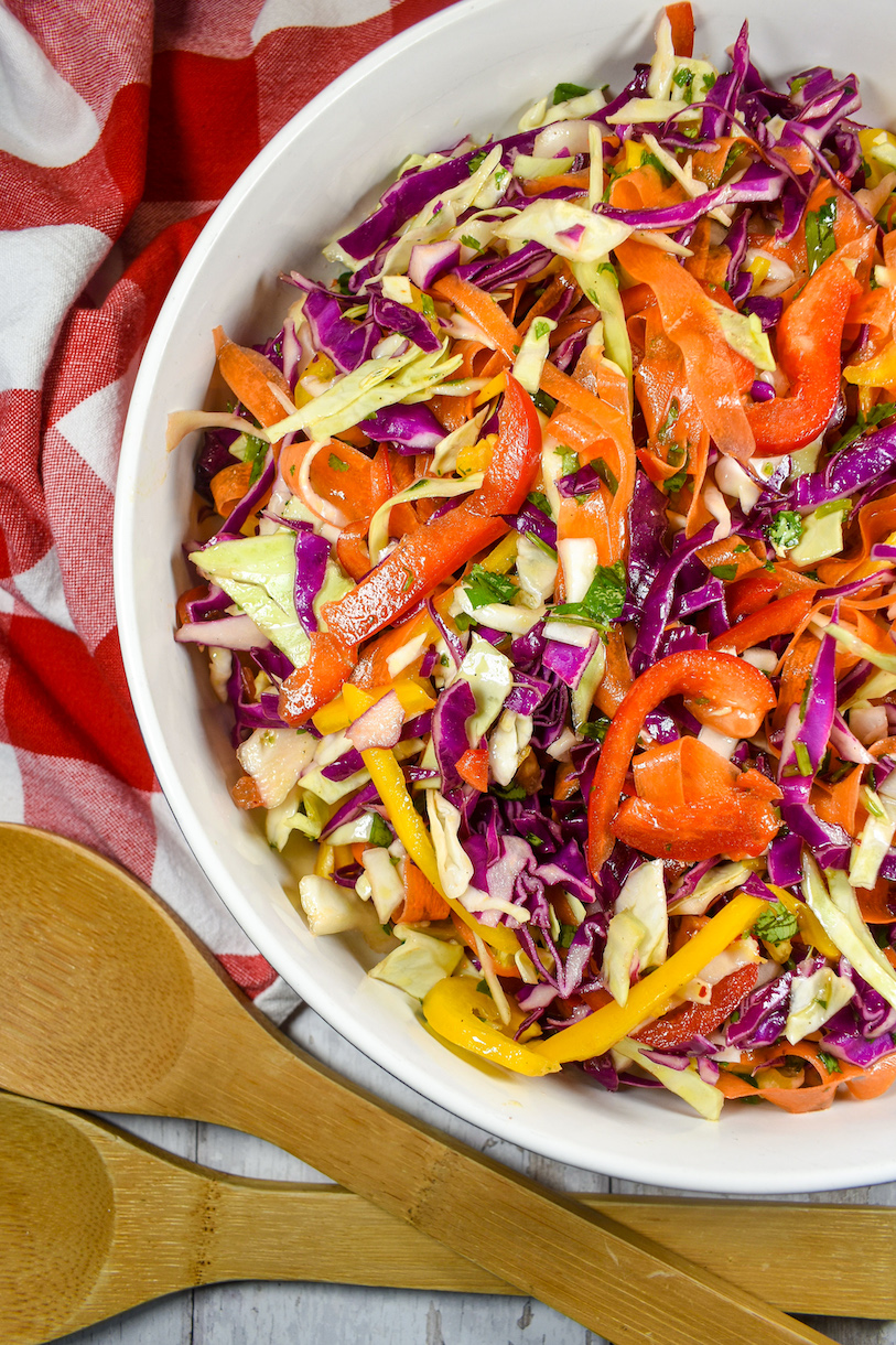 a bowl of rainbow coleslaw with serving spoons and a red checkered tea towel