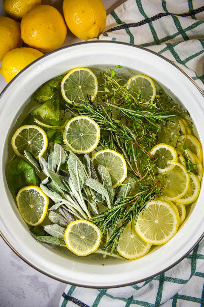 looking down at a mediterranean herb simmer pot, arranged on a white surface with a plaid towel and yellow lemons