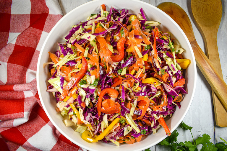 a bowl of rainbow coleslaw, wooden serving spoons, and a red gingham towel