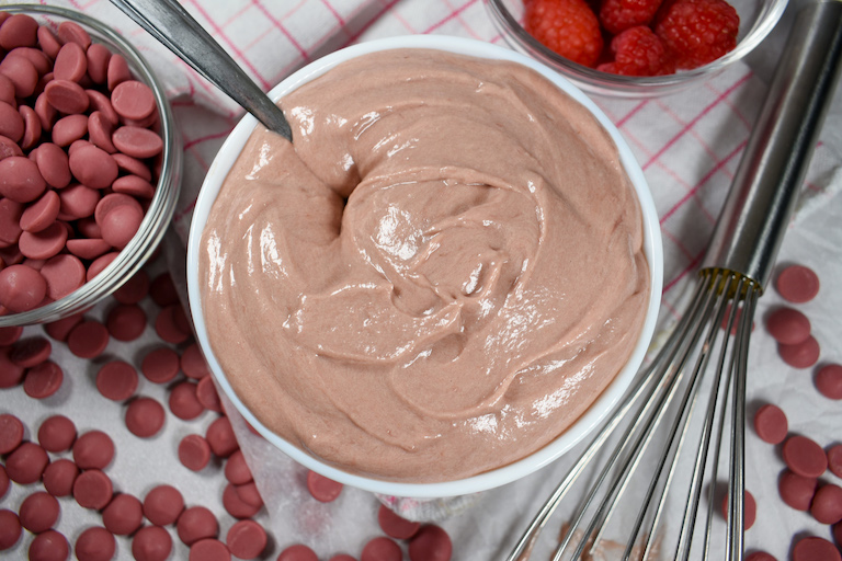 a bowl of ruby chocolate ganache surrounded by raspberries, ruby callets, and a wire whisk