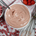 bowl of ruby chocolate ganache with ruby callets and a plaid towel, a bowl of raspberries, and a whisk