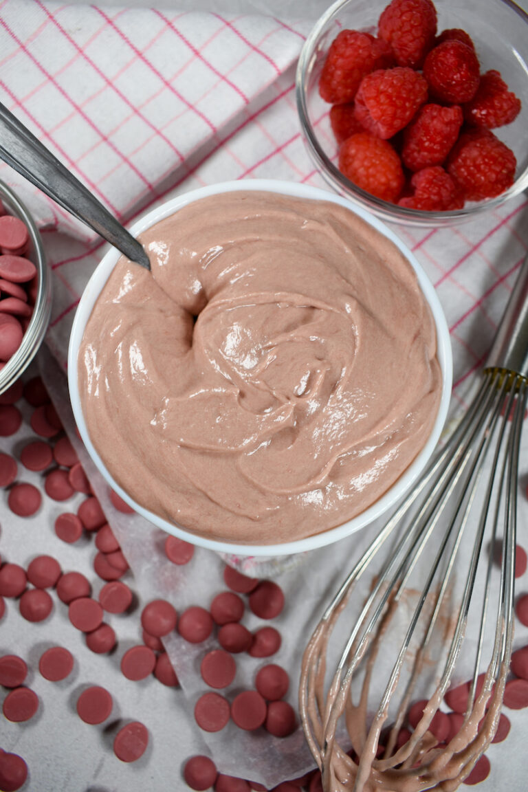 bowl of ruby chocolate ganache with ruby callets and a plaid towel, a bowl of raspberries, and a whisk