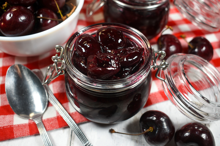 a jar of cherry compote on a red gingham towel with spoons and a dish of fresh cherries