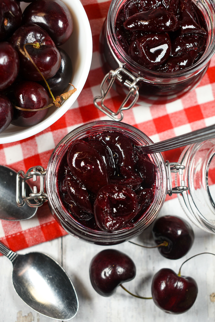 jars of homemade cherry compote, metal spoons, and a dish of fresh cherries arranged on a red and white checkered towel