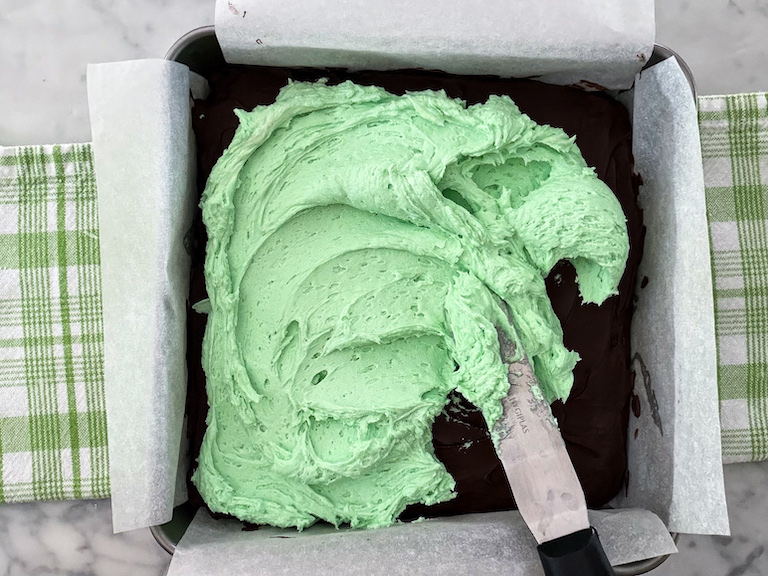 Spatula spreading mint green buttercream over a pan of brownies