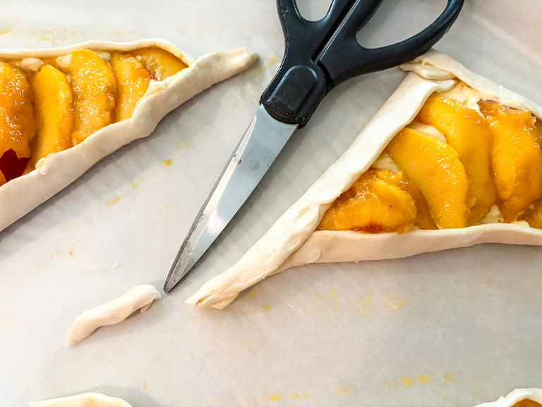 kitchen shears trimming the edges of peach puff pastry triangles