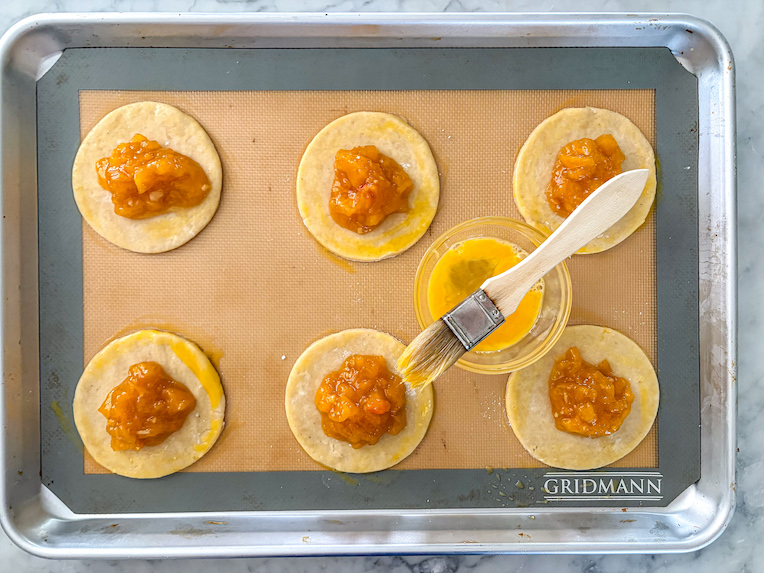 Discs of pastry with peach jam in the middle, on a tray with a dish of egg wash and a pastry brush