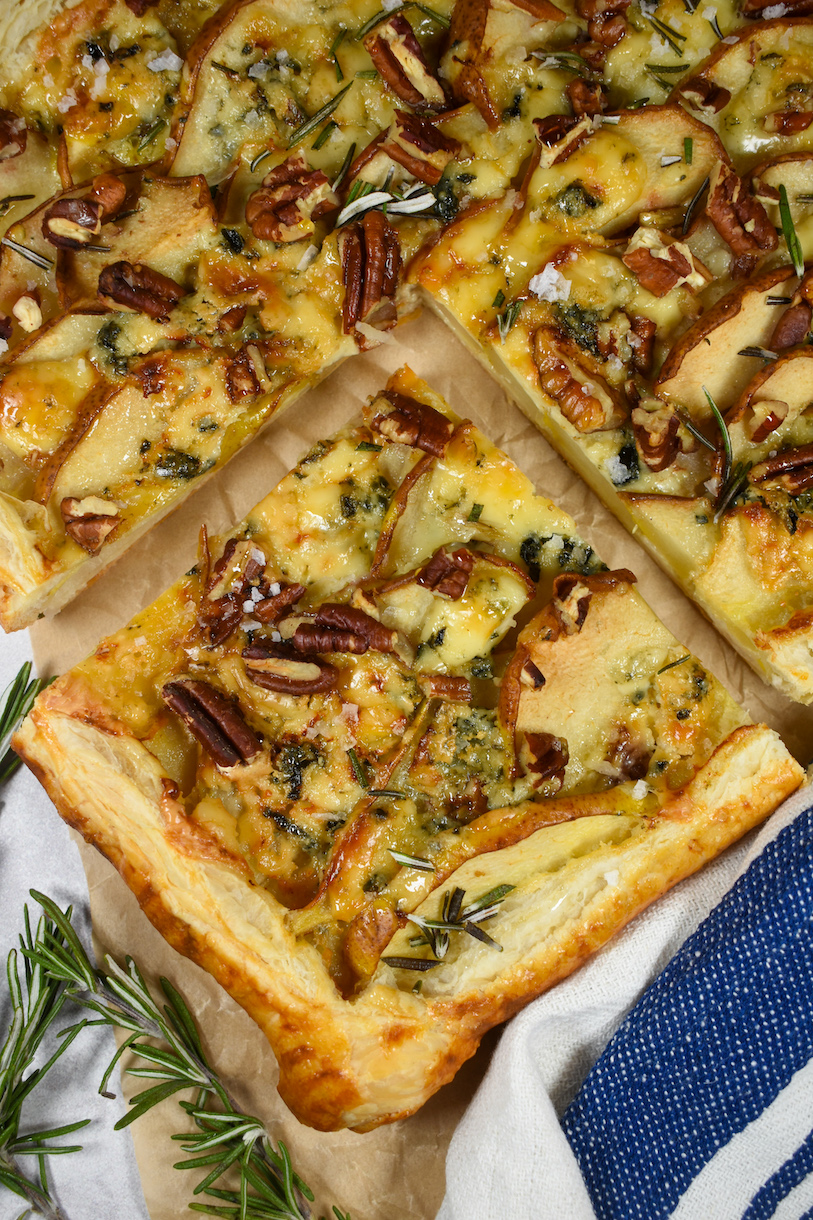 a square slice of pear and blue cheese tart with a striped towel and sprigs of rosemary
